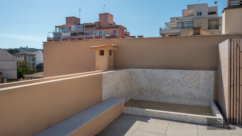 A modern rooftop terrace featuring a tiled plunge pool, beige stone flooring, and views of city buildings under a blue sky.