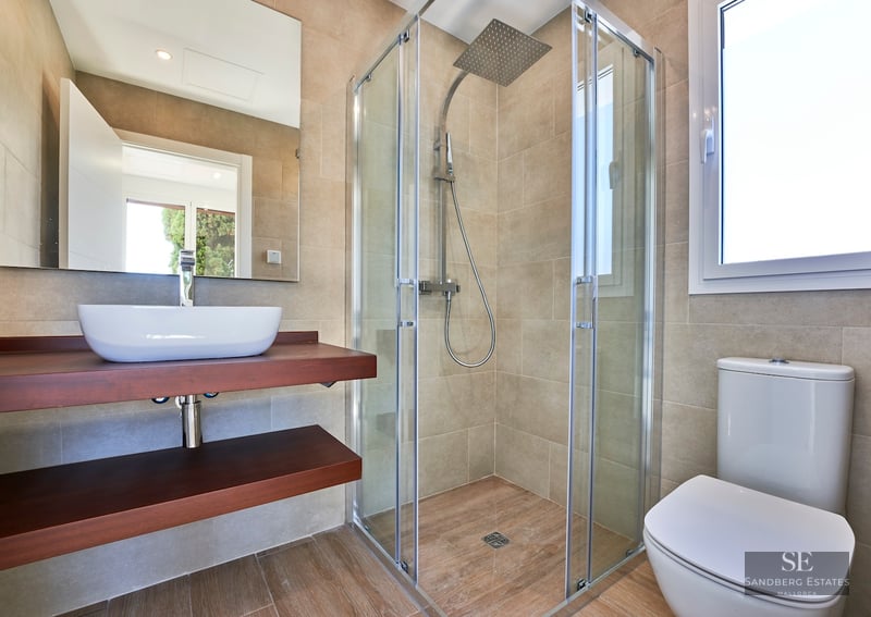 Modern bathroom with beige tiles, glass walk-in shower, wooden floating vanity, vessel sink, and white toilet.