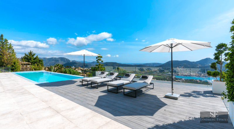 Infinity pool on a wooden deck with grey loungers, white umbrellas, and a vast view of mountains and the sea.