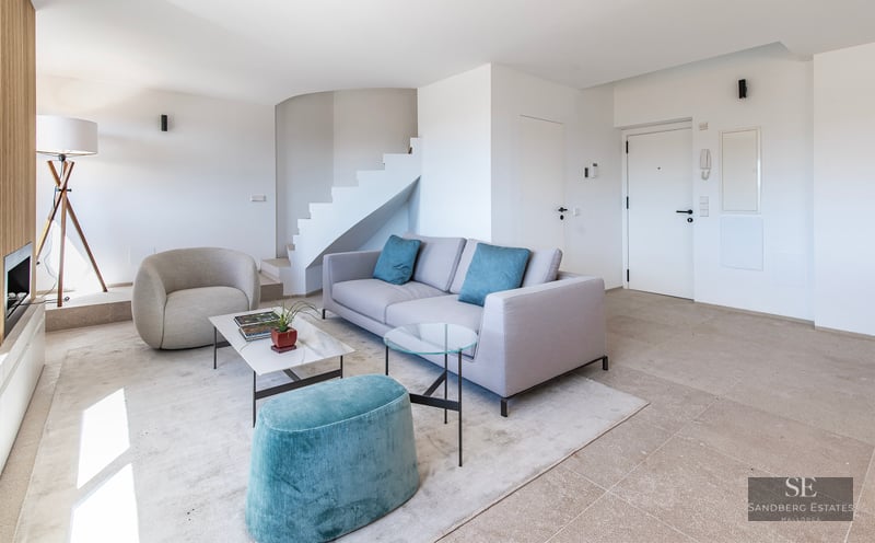Bright minimalist living room with grey sofa, teal ottoman, white staircase, and marble coffee table.
