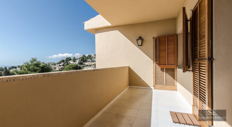 Covered balcony with wooden shutters and tiled floor overlooking the sea and lush greenery.