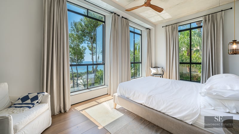 Bright bedroom featuring a white bed, wood floors, a wooden ceiling fan, and large windows with views of the sea and trees.