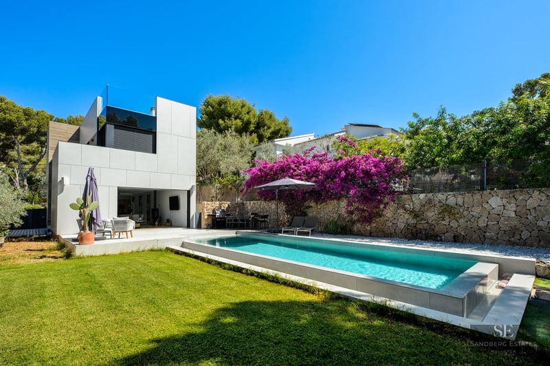 Modern rectangular swimming pool next to a white villa with purple bougainvillea and green lawn under a clear blue sky.