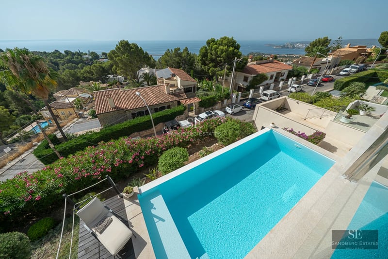 View of a modern infinity pool, surrounded by greenery and with a contemporary house in the background.
