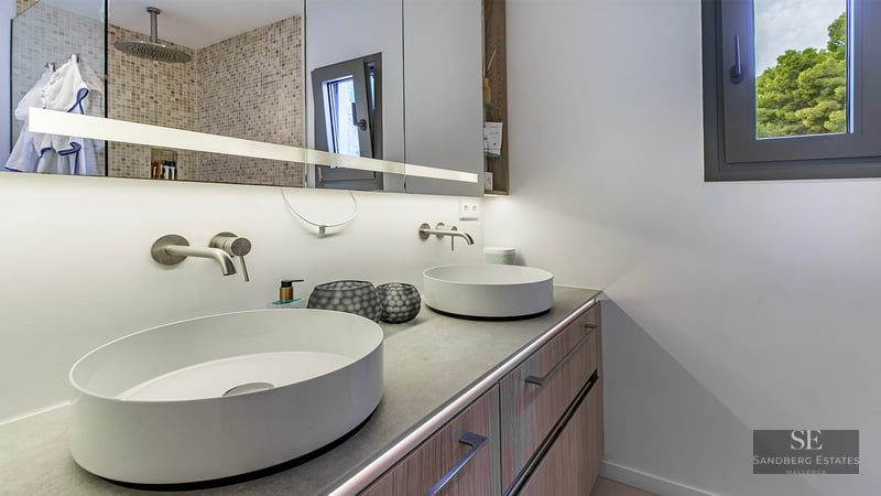 Modern bathroom with two white round vessel sinks on a grey countertop, wood cabinets, and large lit mirror.