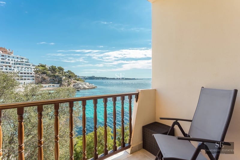 View from a balcony with a wooden railing and grey chair looking over turquoise Mediterranean waters and white buildings.