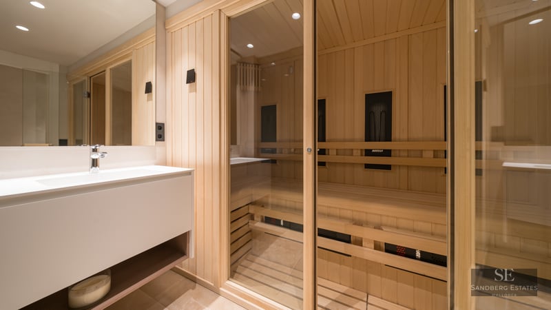 Light wood sauna with glass doors next to a sleek white bathroom vanity and mirror.