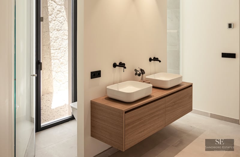 Two white vessel sinks on a floating wood vanity with matte black wall faucets in a bright modern bathroom.