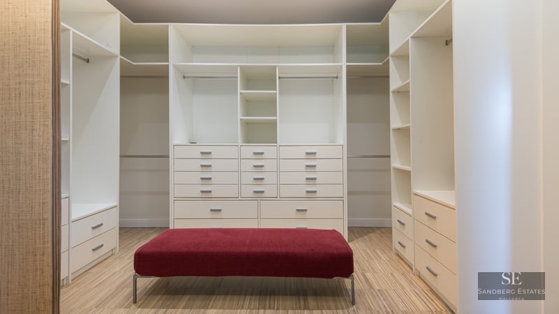Spacious white walk-in closet with numerous shelves, drawers, wooden flooring, and a central red velvet ottoman.