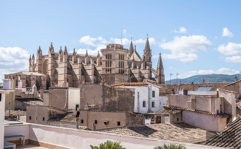 Une grande cathédrale gothique surplombe les toits en terre cuite d'une ville méditerranéenne sous un ciel bleu.