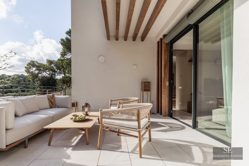 A sunlit modern terrace with a white sofa, wooden furniture, and views of lush green trees through glass doors.