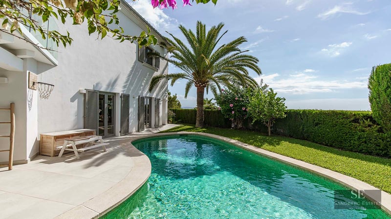 A curved turquoise pool bordered by a lush lawn, a palm tree, and a white villa with grey shutters under a blue sky.