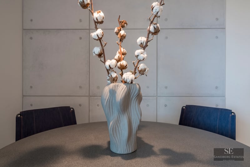 Textured gray vase with cotton branches on a dark table in front of a concrete panel wall.