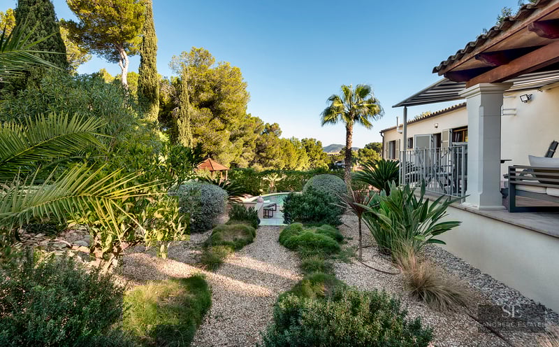 Lush landscaped garden with gravel paths leading to a swimming pool, next to a Mediterranean style villa.