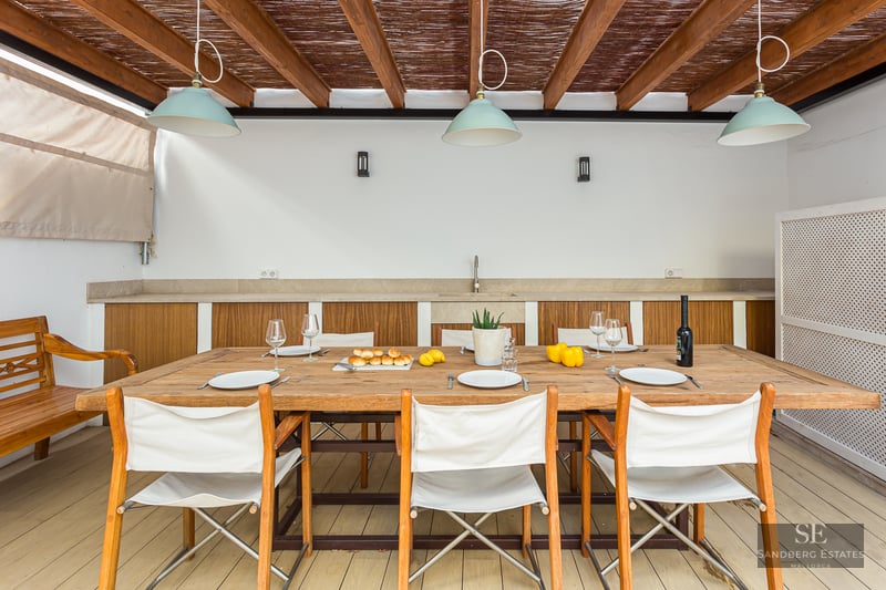 Covered terrace with large wooden table, white canvas chairs, and an outdoor kitchenette under a wooden pergola.