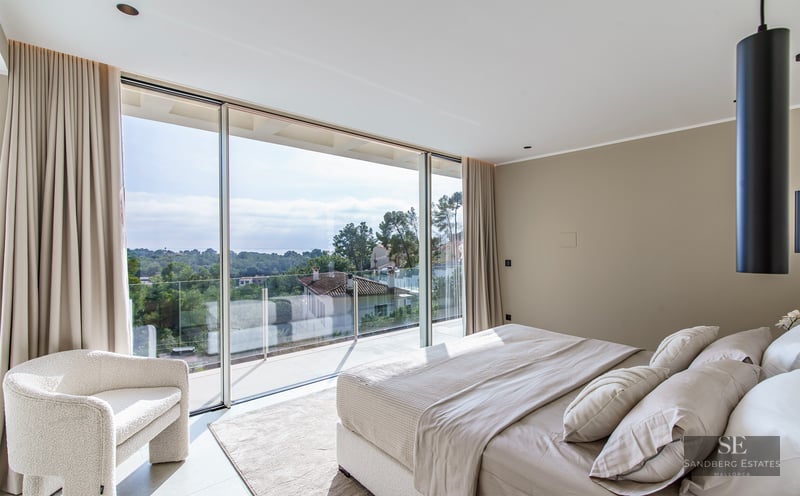 Luxury bedroom with floor-to-ceiling glass doors overlooking trees and houses, featuring a plush bed and boucle chair.