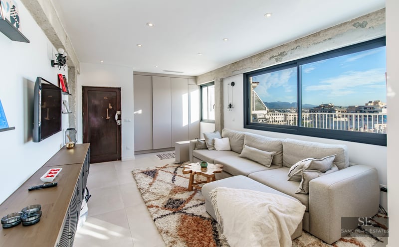 Bright living room with a large grey sofa, exposed concrete beams, and a wide window overlooking a city skyline.