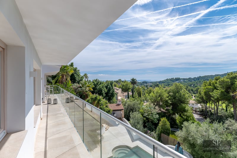 Balcony with glass railing overlooking lush green trees, a swimming pool, and the Mediterranean sea.