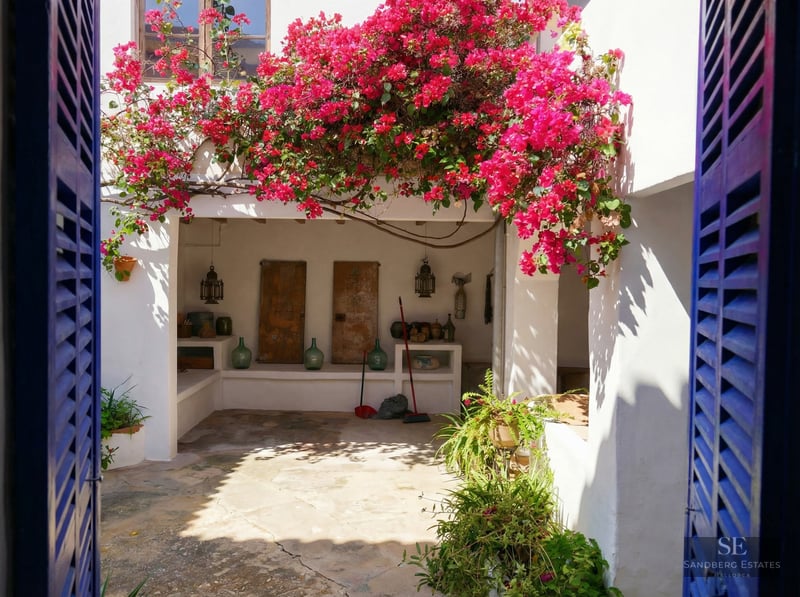 A sun-drenched white courtyard framed by blue shutters, featuring vibrant pink bougainvillea and rustic stone flooring.