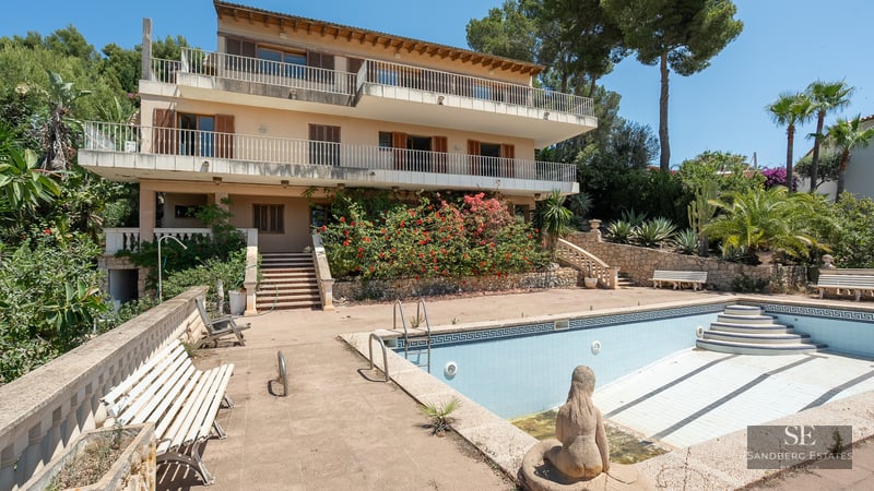Multi-level Mediterranean villa featuring large terraces and a large empty swimming pool surrounded by lush greenery.
