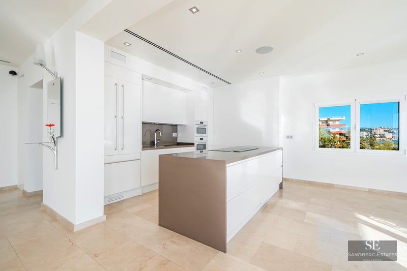 Bright, white modern kitchen featuring a central island, integrated appliances, and sea views through the window.