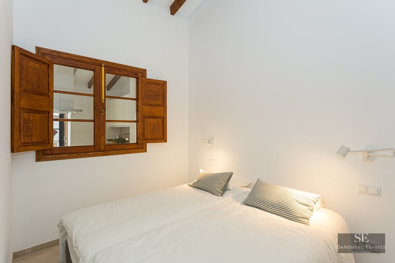 Bright bedroom with white bedding, striped pillows, a traditional wooden window with shutters, and exposed beams.