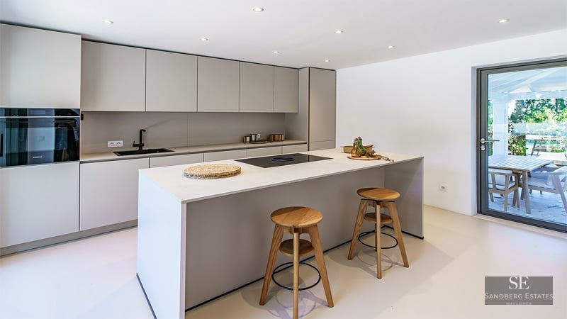 Sleek light grey kitchen with a large island, wooden stools, and glass door leading to a terrace.