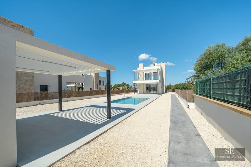 Exterior of a modern villa featuring a blue rectangular pool, white pergola, and gravel courtyard under a clear sky.