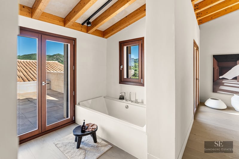 White built-in bathtub under a wooden beam ceiling with access to a terrace and mountain views.