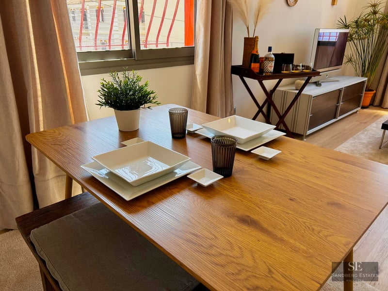 Wooden dining table set with white square plates and glasses in a bright living room.