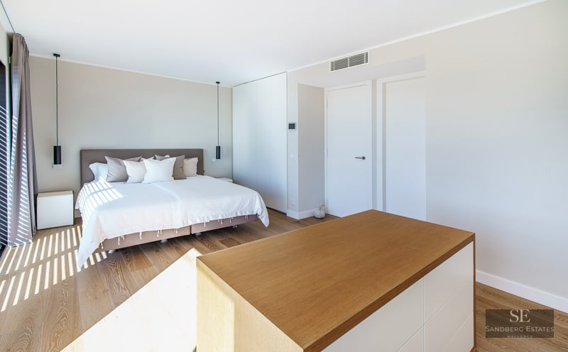 Modern bedroom with a large bed, light wood floors, and natural sunlight casting linear shadows.