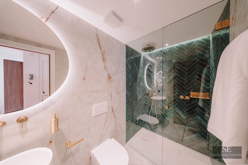 Modern bathroom with white marble walls, a round backlit mirror, and a walk-in shower with green herringbone tiles.