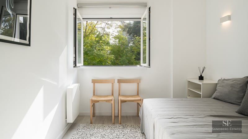 Bright minimalist bedroom with white walls, wooden chairs, grey bed, and an open window viewing lush green trees.
