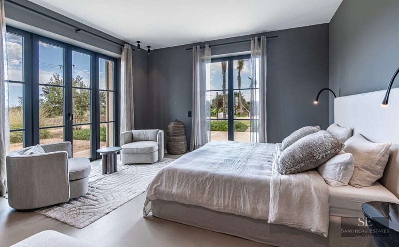 Elegant bedroom with grey walls, a large bed, armchairs, and floor-to-ceiling windows opening to a garden.