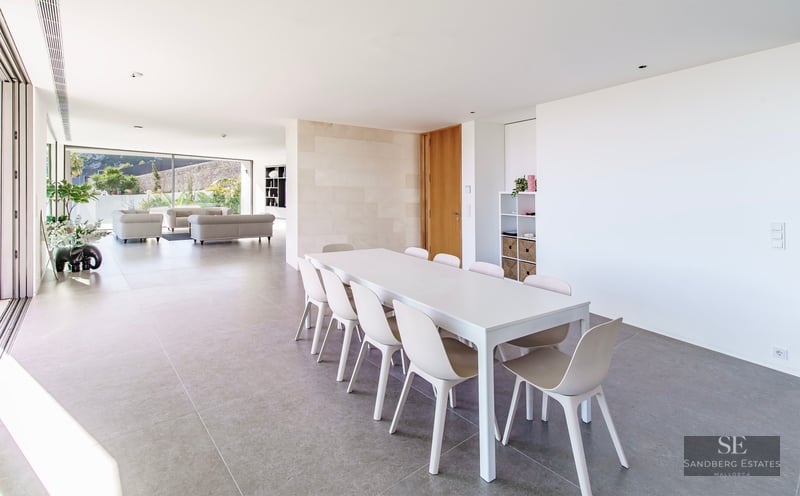 Bright minimalist dining room with white table and chairs, leading into a spacious living area with floor-to-ceiling windows.