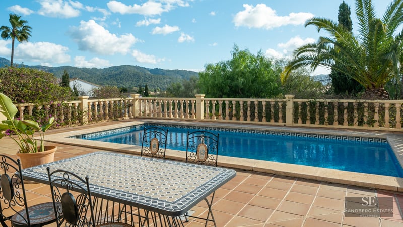 Rectangular swimming pool and mosaic dining table on a terracotta terrace with mountain views and palm trees.