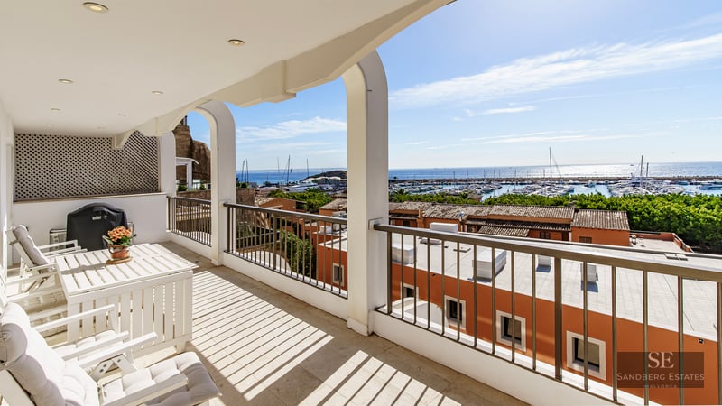 White arched balcony with outdoor furniture overlooking a harbor with boats and blue sea under a clear sky.