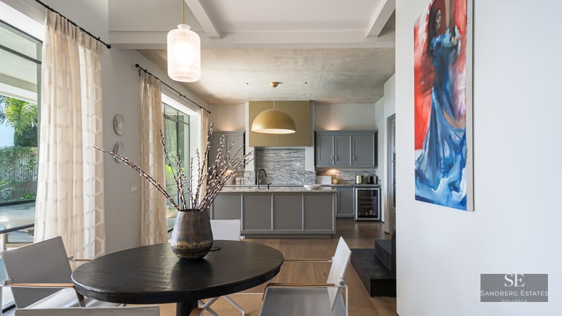 Modern kitchen with grey island, marble backsplash, gold lamp, and dining table with chairs by a large window.