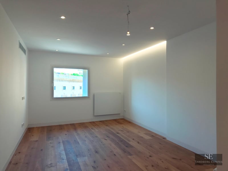 Empty white room with light oak wood floors, recessed ceiling lights, and a window showing a classical building outside.