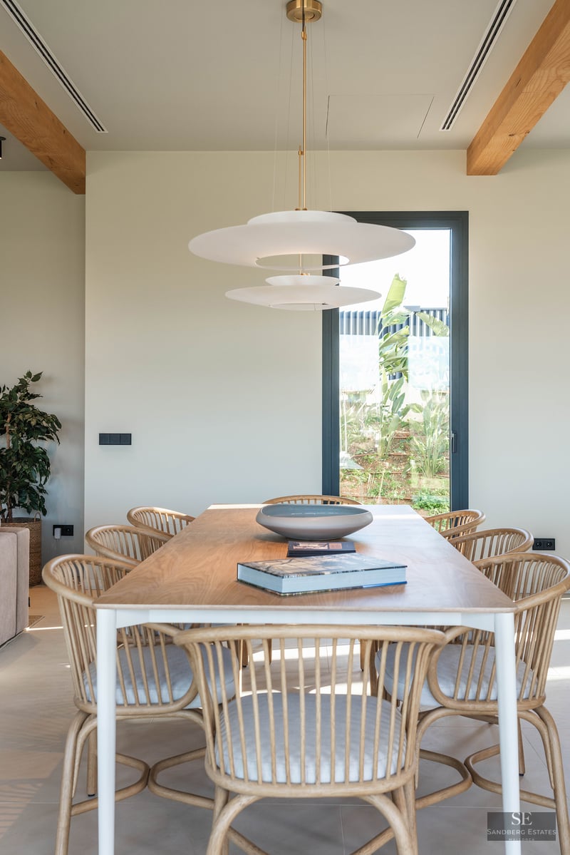 Modern dining room featuring a wooden table, rattan chairs, a designer pendant light, and a window with garden views.