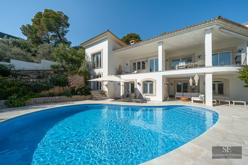 Grande piscine bleue devant une villa méditerranéenne blanche à deux étages avec terrasses et arbres.