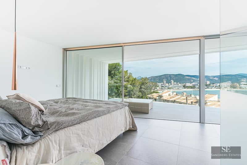 Bright modern bedroom with a large bed and floor-to-ceiling glass doors opening to a terrace with coastal views.