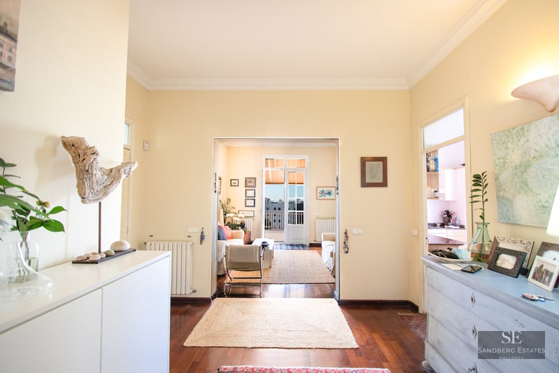 Hallway with wood floors leading to a bright living room with sofas and a door to a sunny outdoor balcony.