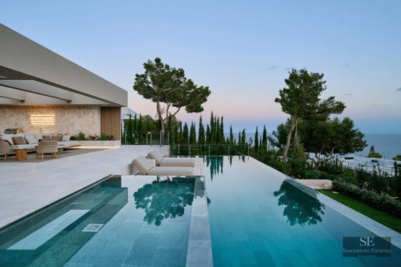 Modern infinity pool and stone terrace with lounge furniture overlooking the ocean at twilight.