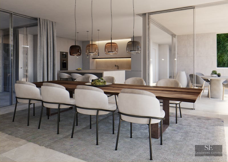 Large wooden dining table with cream chairs and glass pendant lights next to an open terrace and modern kitchen.