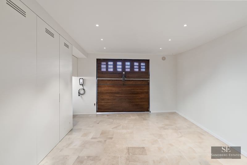 Clean modern garage with beige stone tiles, a dark wood garage door, white cabinets, and an EV charging station.