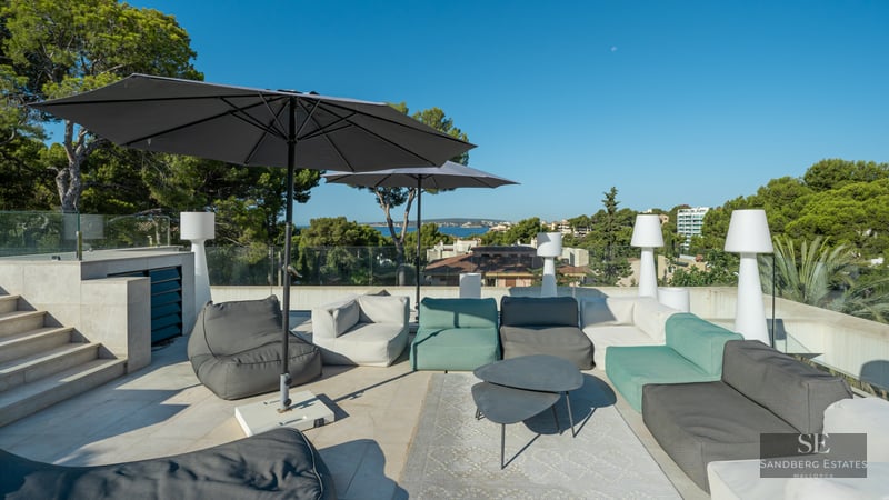 Modern rooftop terrace with grey and green lounge sofas, large black umbrellas, and glass railings overlooking the sea.