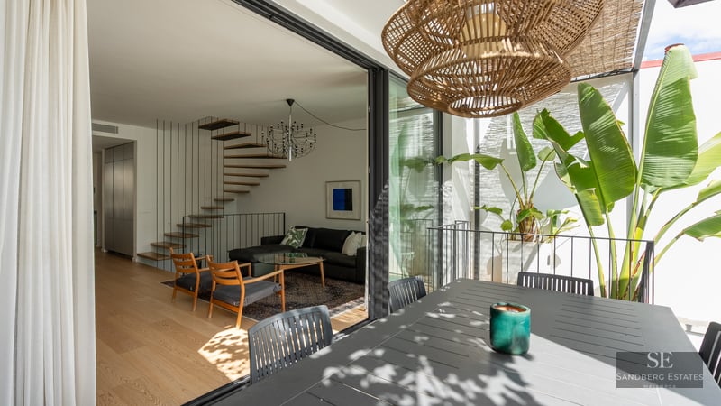 Modern living room with a floating wooden staircase seen through open glass doors from a terrace with tropical plants.