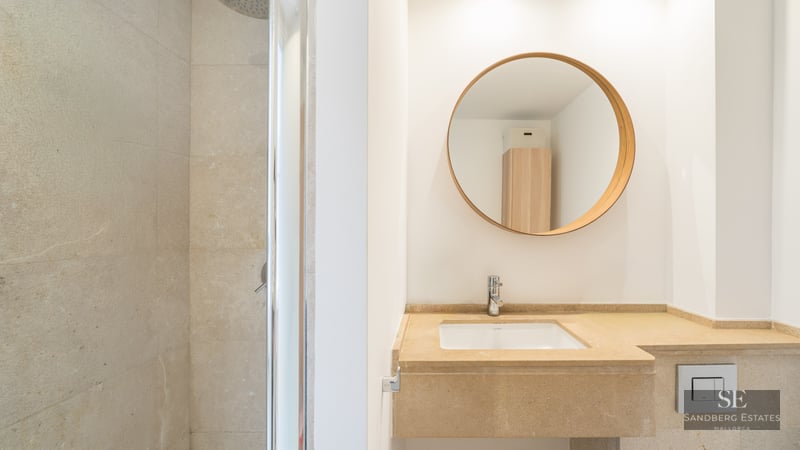 Contemporary bathroom featuring a beige stone vanity, round wooden mirror, and a glass walk-in shower.
