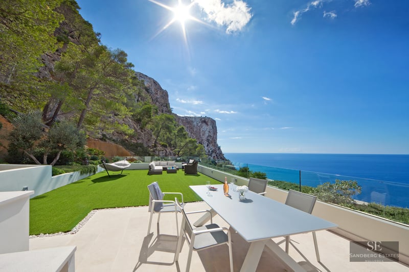 Modern outdoor terrace with dining area, artificial lawn, and glass railing overlooking the sea and cliffs.